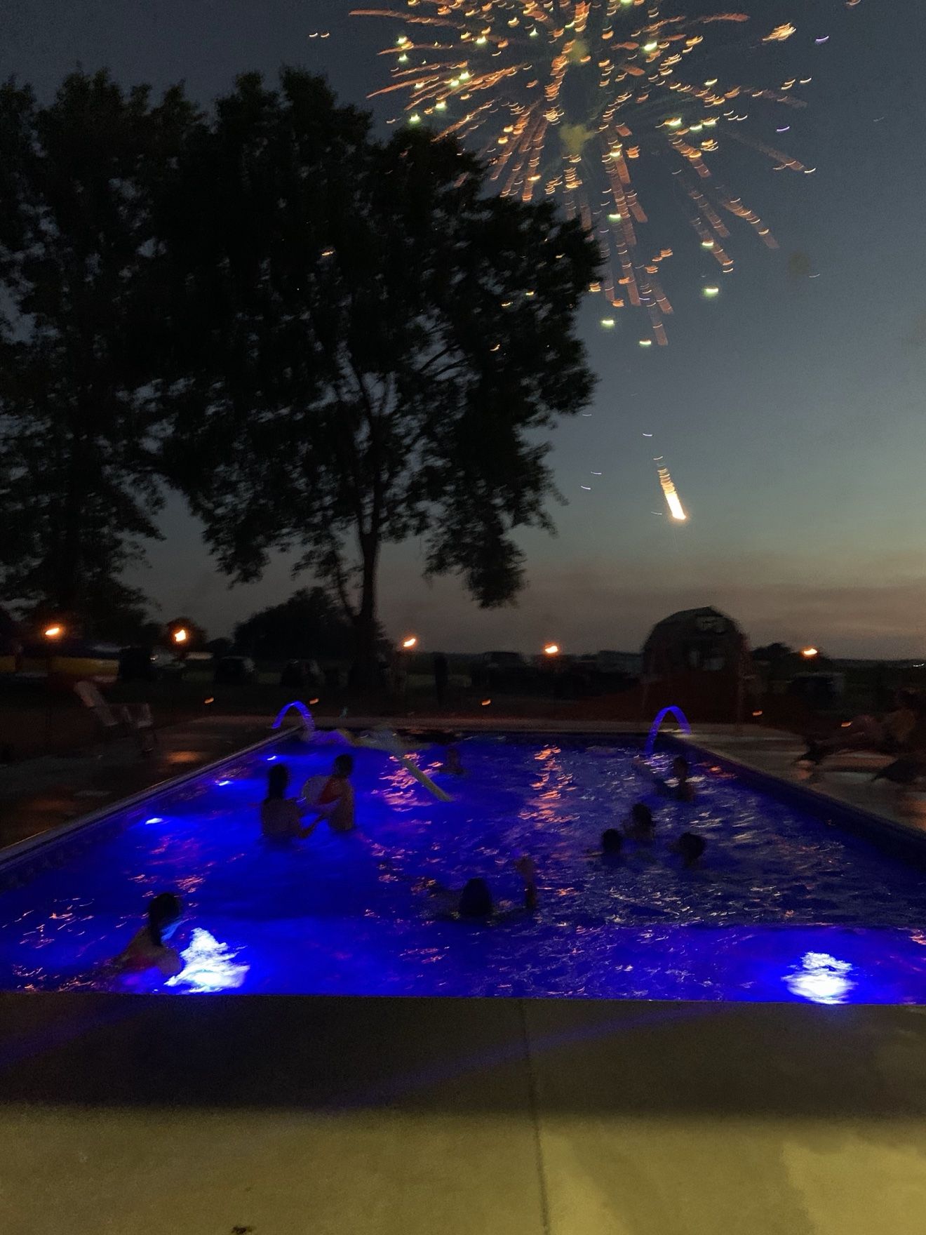 Fireworks bursting over the glowing pool at dusk while kids swim below